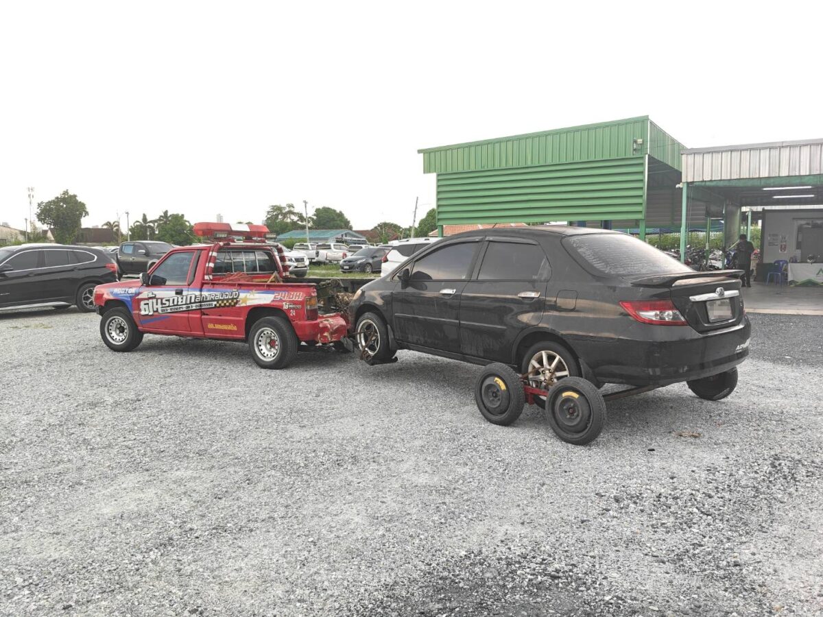 Red tow truck with a trailer hitch towing a black sedan on a wheeled dolly in a gravel lot with a green metal building in the background